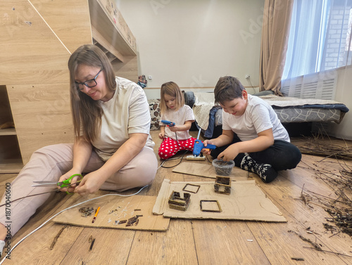 A mother and children sit on the floor and do crafts using wooden sticks, cardboard, scissors, and a glue gun, encouraging creativity and family time. A mother and children are making a craft.