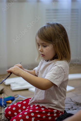 Vertical. A young child is enthusiastically creating an art project using natural tree branches and glue. The child develops creativity and fine motor skills at home while creating the craft.