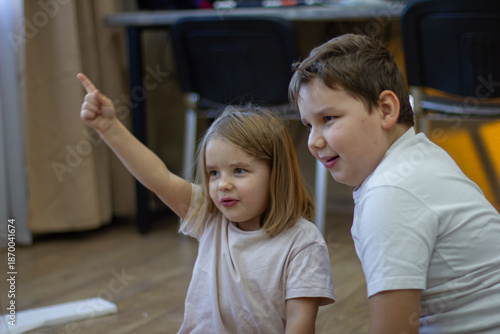 Children joyfully make a craft. Two young siblings sit on the floor at home, sharing a moment of discovery, pointing and smiling with curiosity and wonder as they playfully learn and bond.
