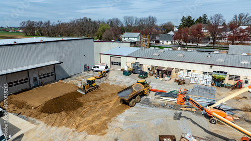 Construction equipment is busy at a site with trucks, loaders, and piles of materials. Workers are moving dirt and supplies in an area near buildings and trees under a clear sky.