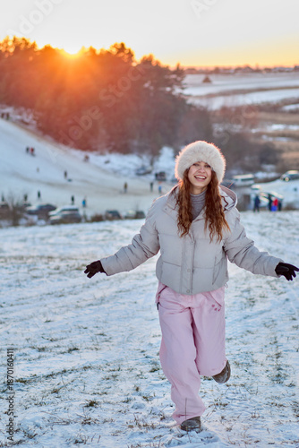 A smiling young woman in winter clothing runs happily across a snowy field with a backdrop of a snowy hill, forest, and beautiful sunset, showcasing the joy of winter.