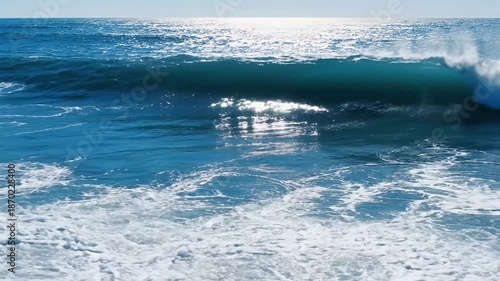 This vibrant ocean footage captures the dynamic beauty of large blue waves as they rhythmically crash, creating mesmerizing white foam and spray along the coastline. The bright sunshine glimmers acros