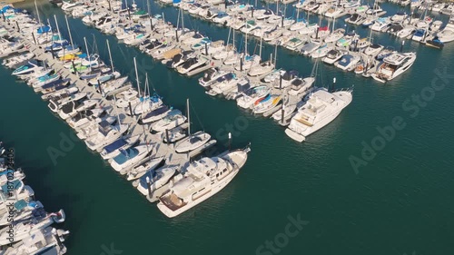 Aerial view of a coastal marina filled with docked boats and yachts, surrounded by waterfront roads, green parks, and hillside neighborhoods under clear daylight skies.