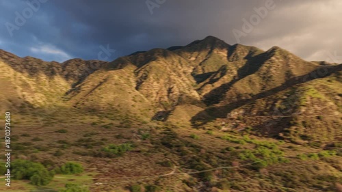 Aerial view of rugged mountain slopes illuminated by warm sunlight, with dramatic shadows, textured ridgelines, and patches of green vegetation beneath a moody sky.