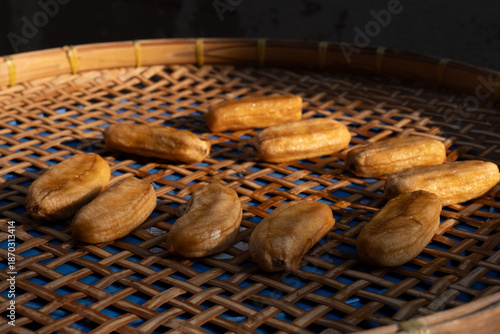 Perspective view of sun-dried bananas arranged on a woven bamboo tray, illuminated by sunlight against a dark background.