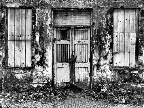 Facade of an entrance of an old abandoned budling with mosaic pattern tiles, rectangular window frame with metal sheet. a damaged wooden door with zinc covering the broken parts.