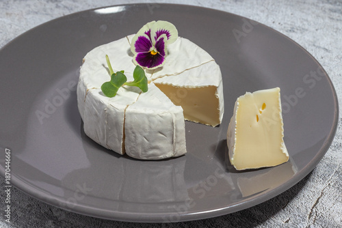 Close-up of a creamy Brie wheel with a slice cut out, garnished with a vibrant edible flower and microgreens on a gray plate.