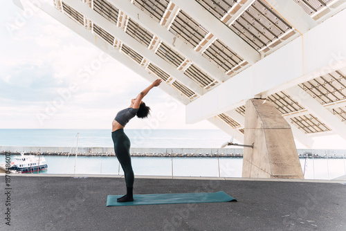 Fit woman performing yoga upward salute pose under a futuristic white metal structure by the ocean