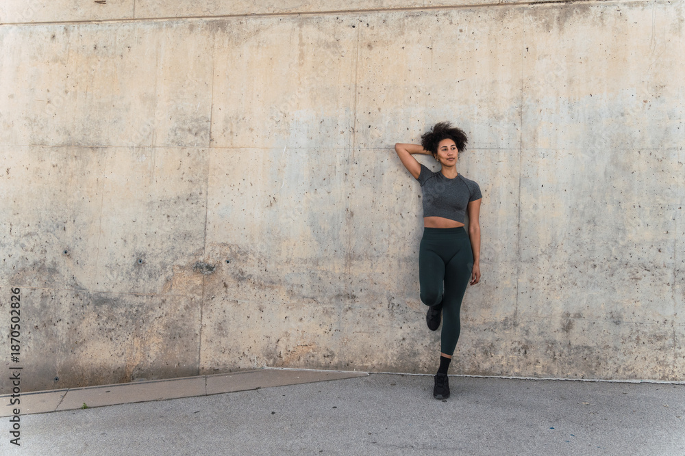 Fototapeta premium Sporty woman with curly hair and sportswear leaning against concrete wall after outdoor workout