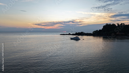 White luxury yacht rocking gently on calm ocean waters near a tropical island, with vibrant sunset hues of orange and pink reflecting on the tranquil sea