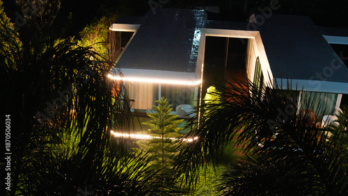 Warm lights glowing around a modern tropical bungalow at night create a cozy, inviting atmosphere, nestled among lush foliage and surrounded by serene greenery