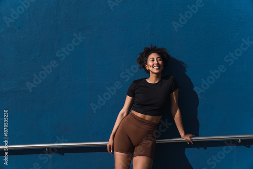 Smiling young woman with brown shorts and black t-shirt leaning on a railing in front of a blue wall
