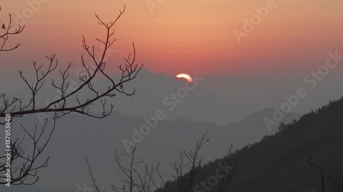 4K fast motion footage of sunrise viewed from top of mountain, sun rising behind mountains far away, golden sun with orange sky background and silhouette of tree branch foreground, peaceful landscape