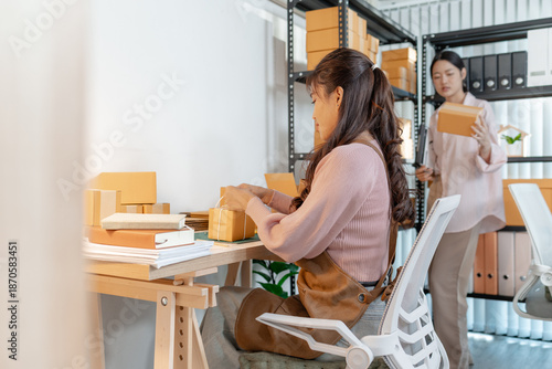 Two young women working together in a small business office, preparing cardboard boxes, checking inventory, and handling packaging tasks as part of their startup online store operations.