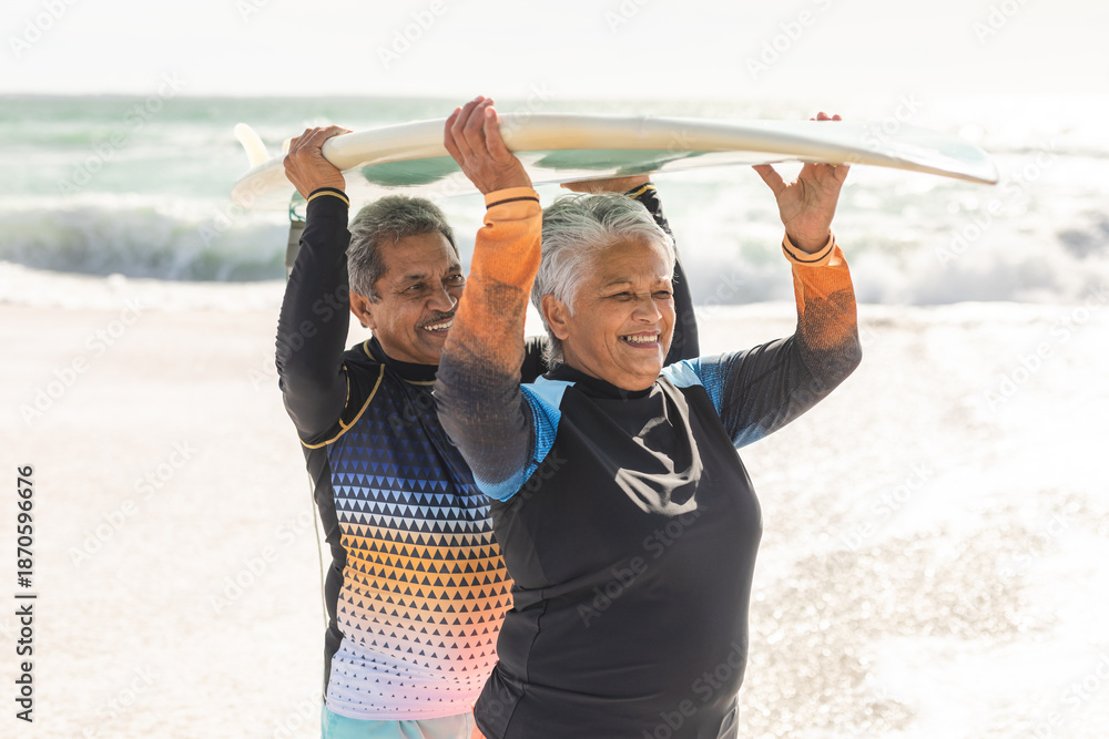 Naklejka premium Happy multiracial senior couple carrying surfboard on heads against sea at beach during sunny day