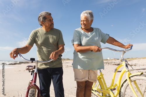 Multiracial senior couple looking at each other standing with bicycles on sunny beach against sky