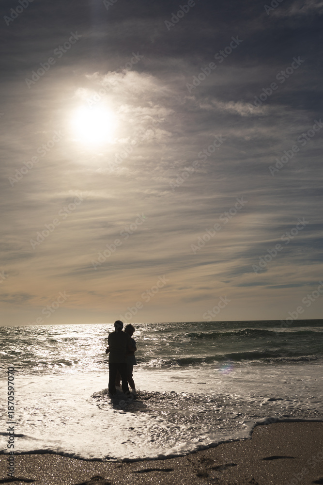 Fototapeta premium Senior retired multiracial couple enjoying waves together on shore at beach against sky