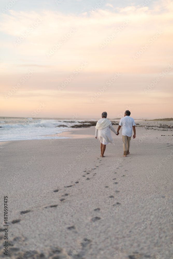 Obraz premium Multiracial senior couple walking while leaving footprints on sand at beach during sunset