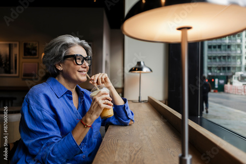 Smiling woman with gray hair drinking juice in urban cafe by window