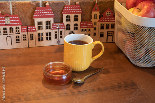 Cozy home tea time with a yellow mug, honey, and a spoon on a wooden table. Whimsical wooden toy houses and a basket of apples create a warm, rustic kitchen atmosphere.