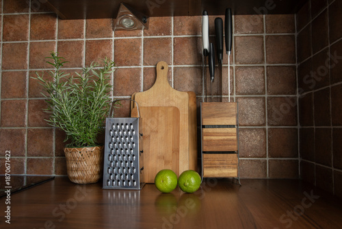 Kitchen counter with wooden cutting boards, knife block, metal grater, fresh rosemary in a pot, and limes. Cozy domestic cooking workspace.
