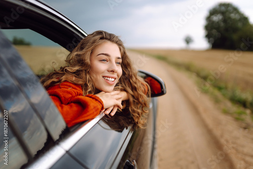 A happy woman leans out the car window, feeling free and enjoying the fresh air at sunset. A traveler on a road trip surrounded by agricultural fields. Concept of travel, freedom, and fun.