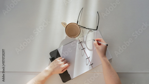 Person's hands writing notes in a planner next to a cup and glasses on a minimalist desk, top view