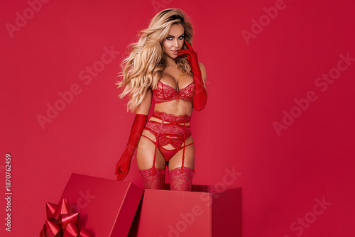 Valentine’s Day fashion portrait of sexy blonde woman in red lingerie standing in gift box with heart balloons on red background, love and holiday concept