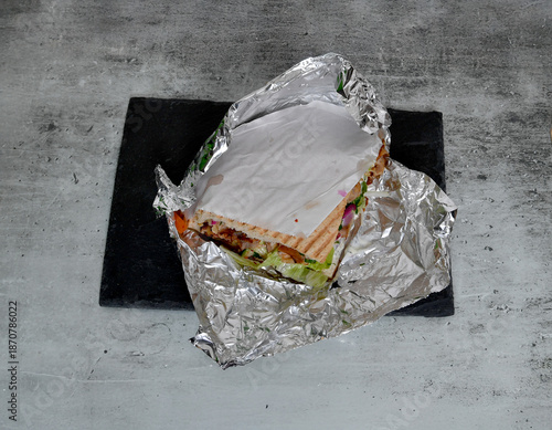 Foto Frisch zubereiteter Döner-Dürüm mit Fleisch, Salat und Sauce, halb in Alufolie gewickelt auf dunkler Unterlage