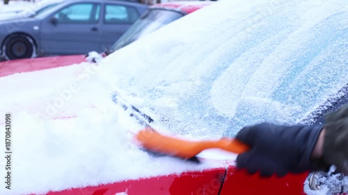 A person scrapes snow off a red car windshield using an orange brush. A gray car is parked in the background. The scene captures winter weather conditions.