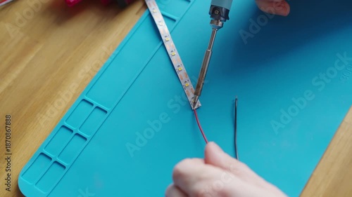 Close-up of male hands soldering red and black wires to an LED strip on a blue silicone mat, detailed electronics repair and DIY technology process