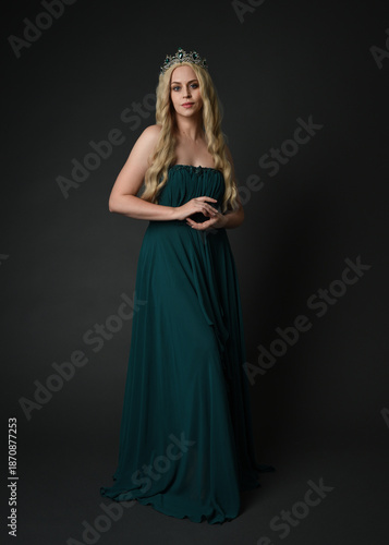 Full length portrait of blonde female model wearing royal green ballgown, fantasy crown. Standing, walking  pose, front view isolated figure on dark studio background.
