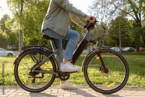Bicyclist rides electric bike in park during sunny day with green trees around in early afternoon