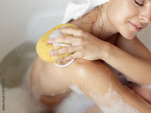 Slika na platnu Close up shot of young happy woman sitting in bathtub with foam taking bath in bathroom rubbing shoulders with sponge