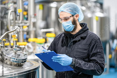 A man in protective gear inspects machinery in a clean industrial setting, taking notes on a clipboard while ensuring safety protocols are followed.