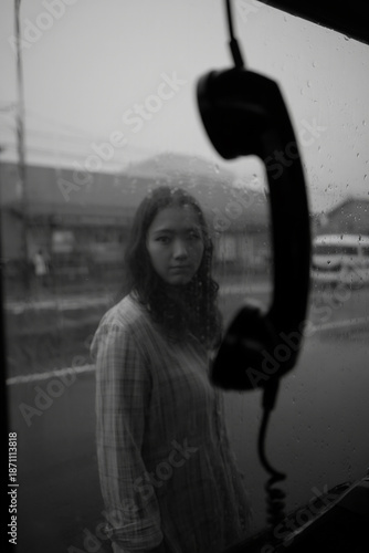 Young woman with a thoughtful expression looks through a rain-streaked window, a blurred telephone receiver in the foreground. A melancholic black and white portrait reflecting urban solitude and refl