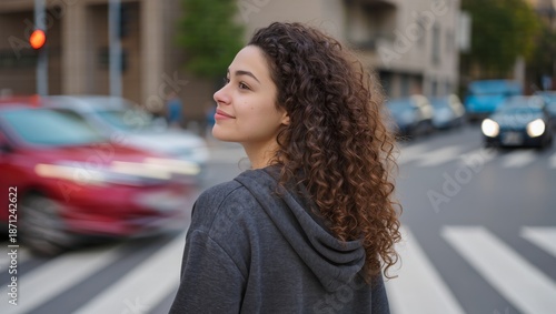 Person in dark gray hoodie stands at pedestrian crossing in city, with blurred cars and buildings behind, red traffic light, capturing urban routine and city movement