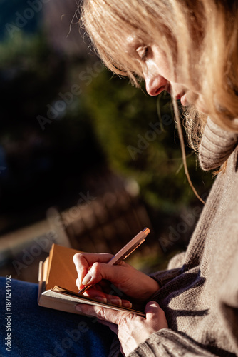 Close up of woman journaling and reflecting on new year intentions on sunny terrace. Mindfulness, slow living, wellbeing, self care, mental health concept. Selective focus on book