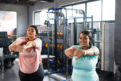 Diverse mid-adult female friends stretching arms forward at gym in pink blue tees near weights
