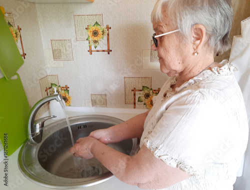 An elderly woman washes her hands with soap under running water from a kitchen sink faucet.