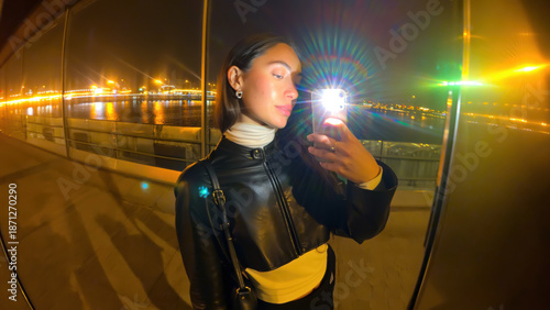 Young woman taking selfie at night with city lights reflecting on water, wearing black leather jacket and white turtleneck, standing outdoors by waterfront, vibrant and confident mood