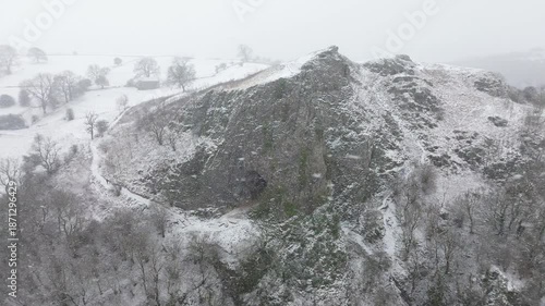 Aerial drone footage panning around Thor's Cave in Derbyshire, England, showing snow covered cave entrance, suitable for travel, tourism, nature and establishing shot use.