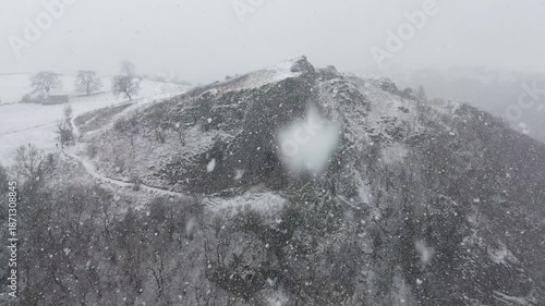 Aerial drone static shot of Thor's Cave in Derbyshire, England, showing snow covered cave entrance during heavy winter snowfall for travel, tourism, nature and establishing shot use.