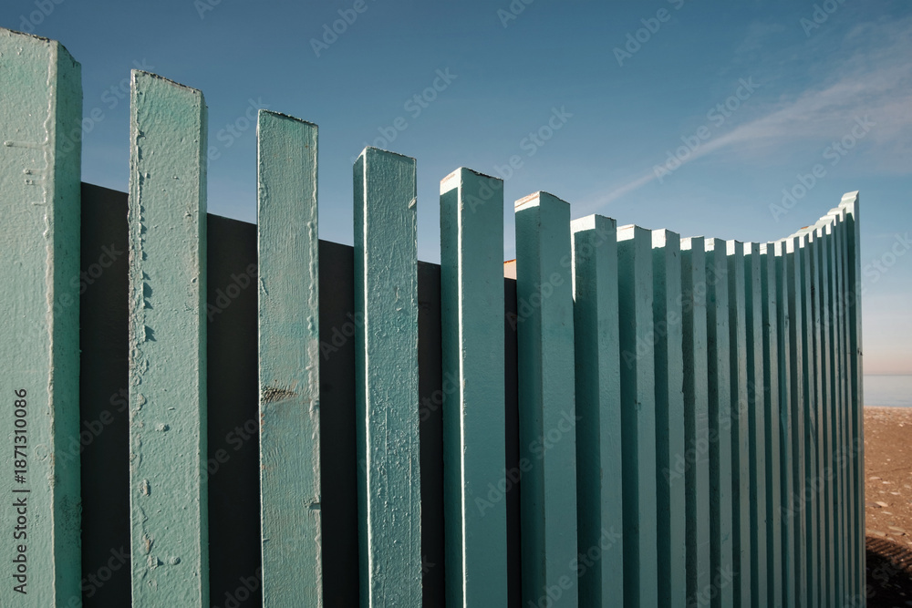 Obraz premium Close-up view of a curved fence made of vertical, closely spaced wooden pales, painted a light blue-green color on blue sky background