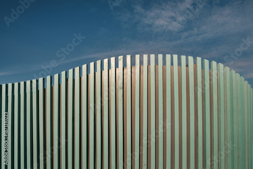 Close-up view of a curved fence made of vertical, closely spaced wooden pales, painted a light blue-green color on blue sky background