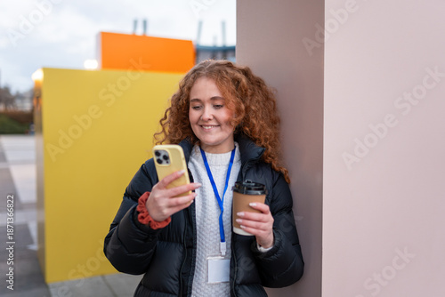 Redhead woman checking smartphone during coffee break outdoors, wearing puffer jacket and lanyard
