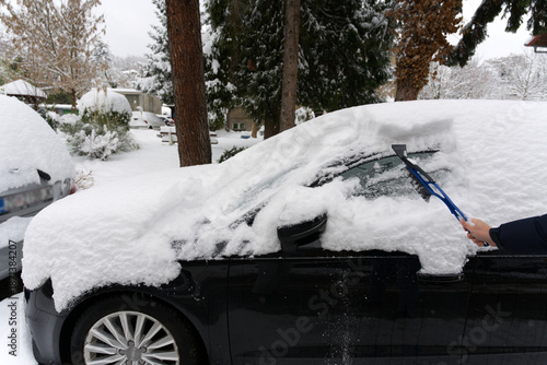 A person clears heavy snow from a black car using a blue brush and scraper, surrounded by snow-covered trees, vehicles, and buildings in a quiet residential neighborhood during a winter storm.