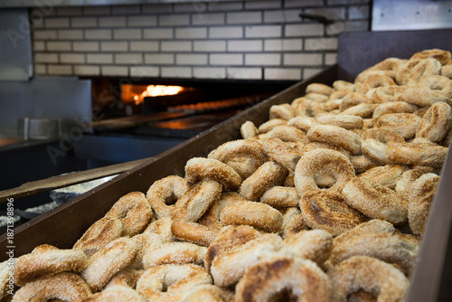 Sesame seed bagels in a metal bin at a bagel shop, oven in the background.