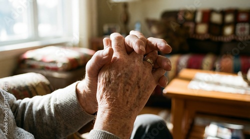 A close-up of elderly hands clasped together, showcasing age lines and a wedding ring, against a cozy living room backdrop.