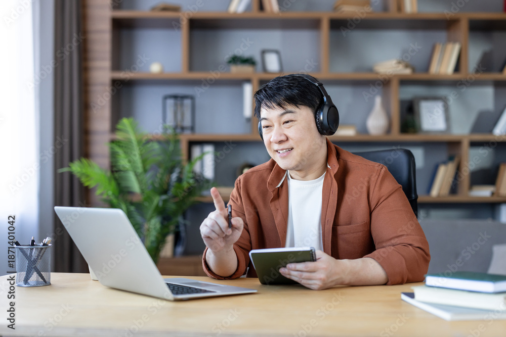 Fototapeta premium Asian man with headphones smiling during a video call from his home office, interacting with laptop and tablet for remote work, e-learning or a virtual meeting, casual smart setup